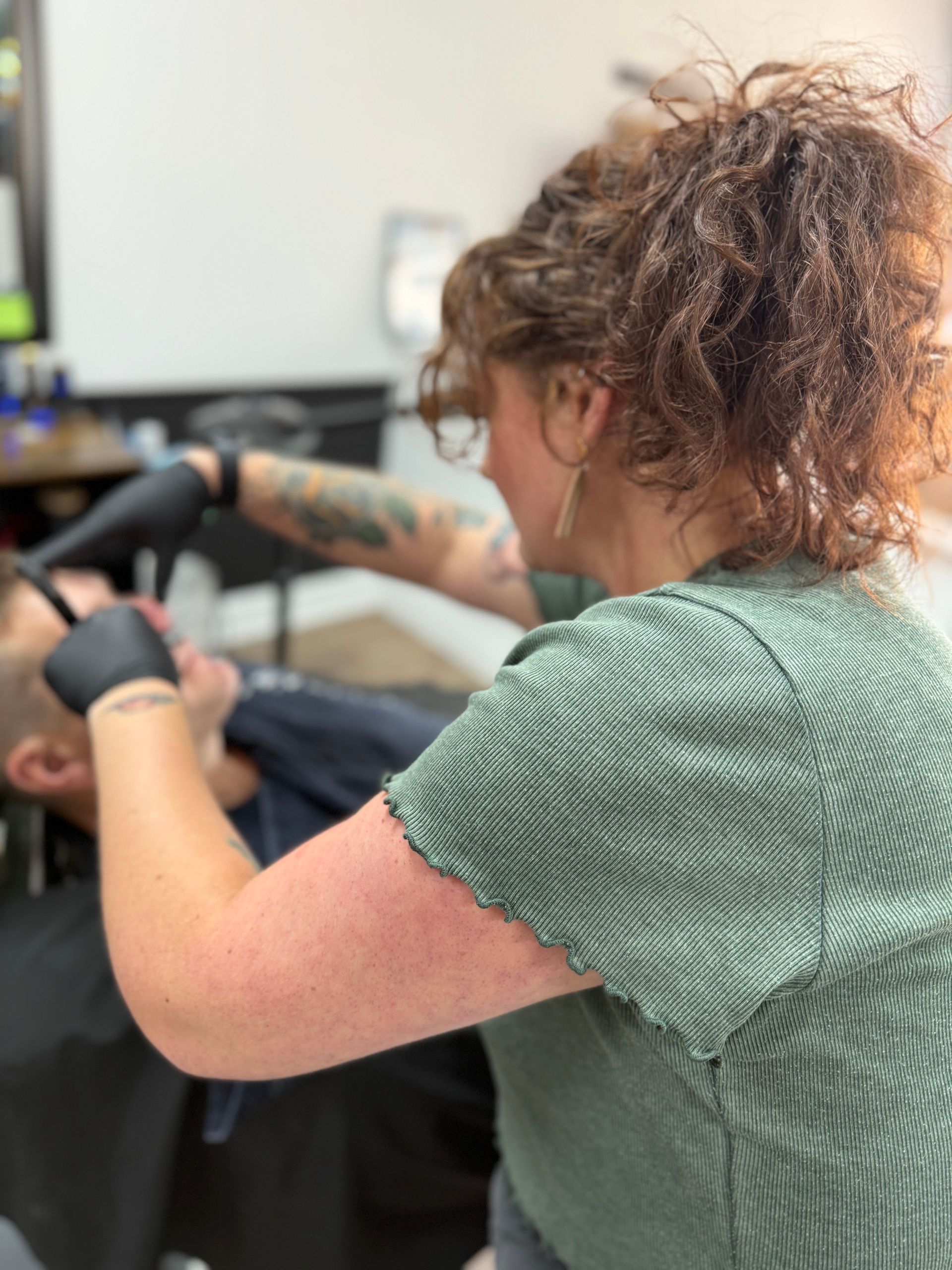 Barber shaving a client's face. The barber has curly hair and wears a green shirt and black gloves.