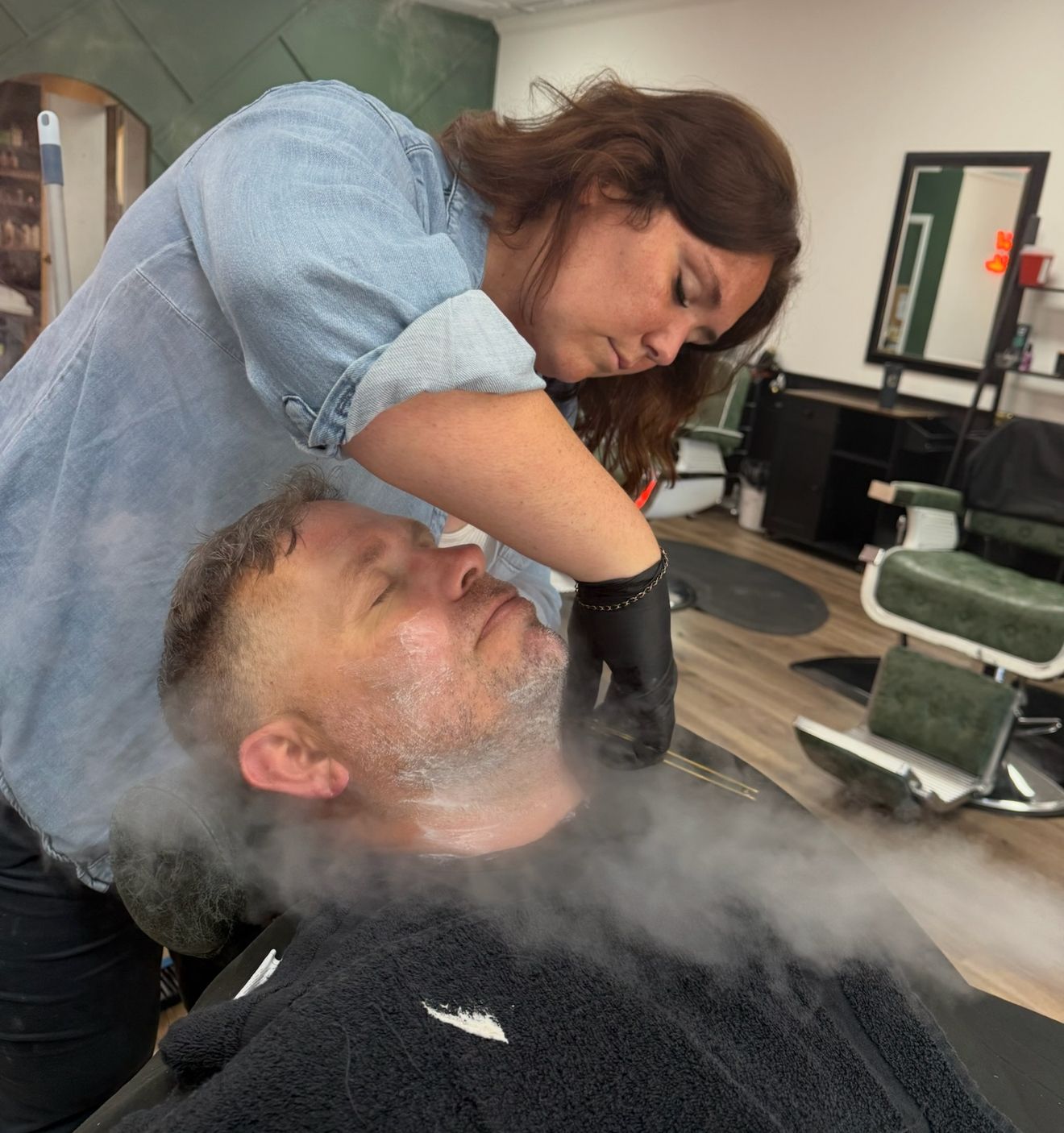 Woman shaves a man's face, steaming. Barber shop setting, black gloves, blue denim shirt, towel.