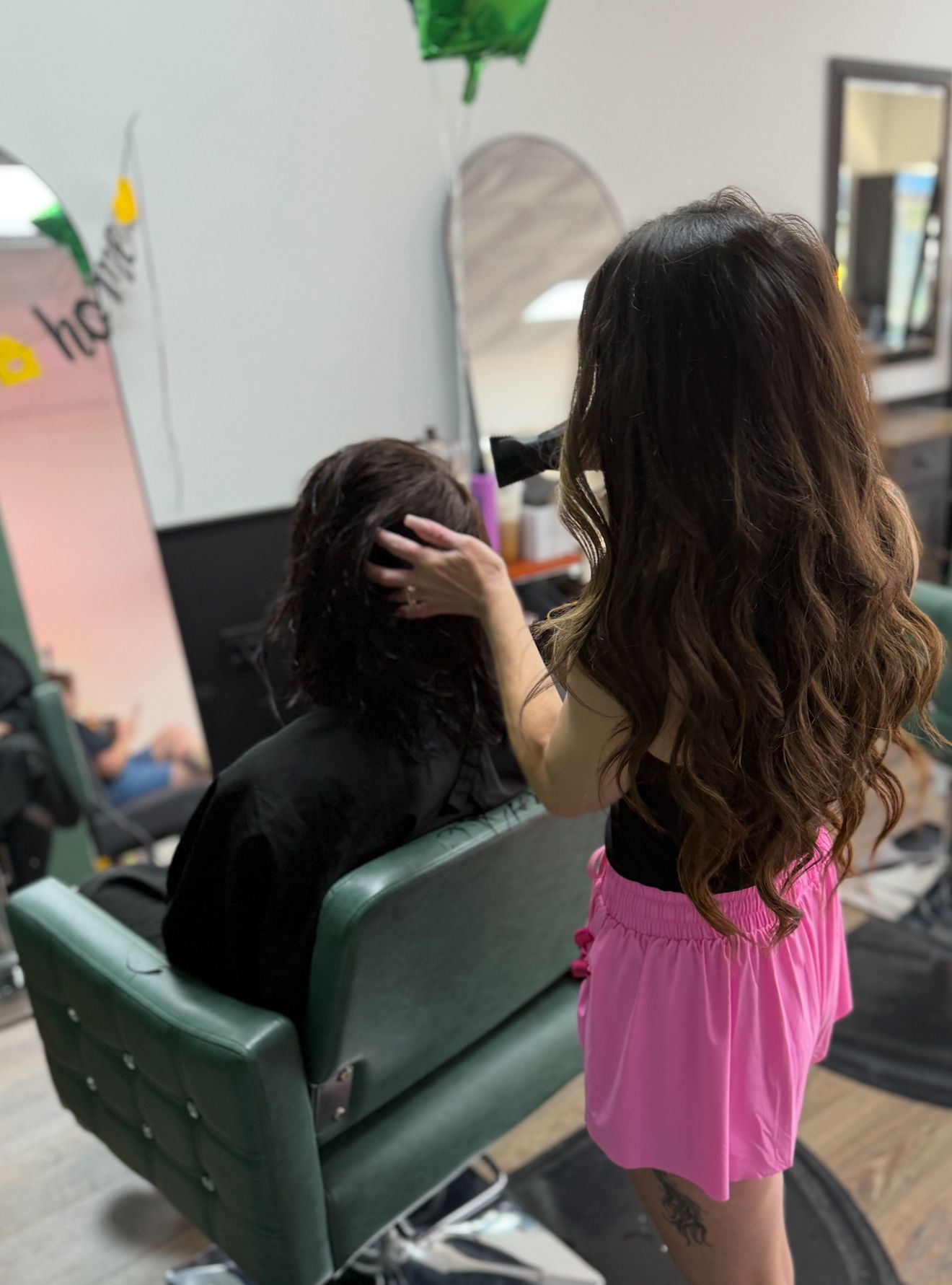 Hairdresser styling a client's hair in a salon. The hairdresser is wearing a pink skirt.