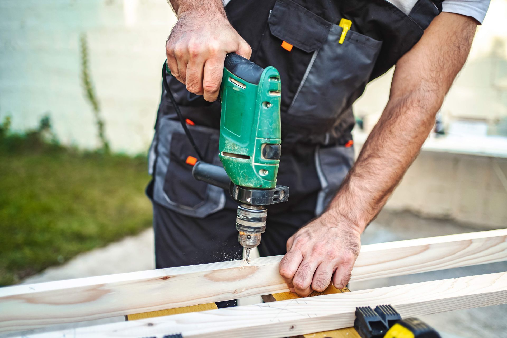 Person using a green power drill to bore a hole into a wooden plank outdoors.