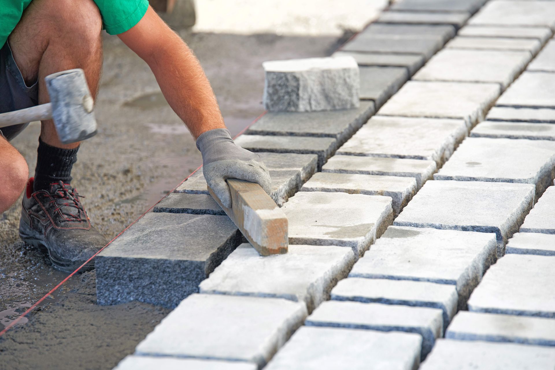 Person laying stone pavers with a hammer and leveling tool on a sandy surface.
