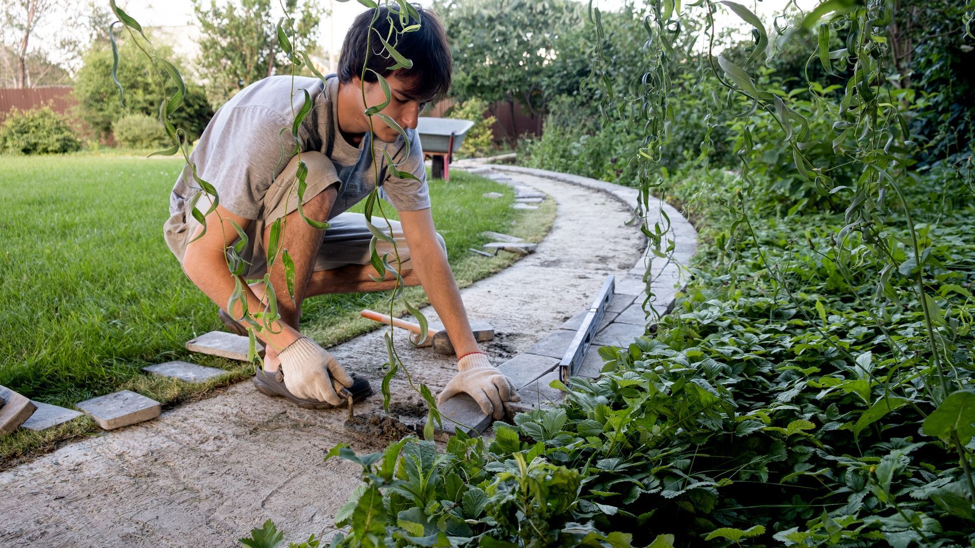 Person gardening, weeding a stone path surrounded by green plants in a yard.