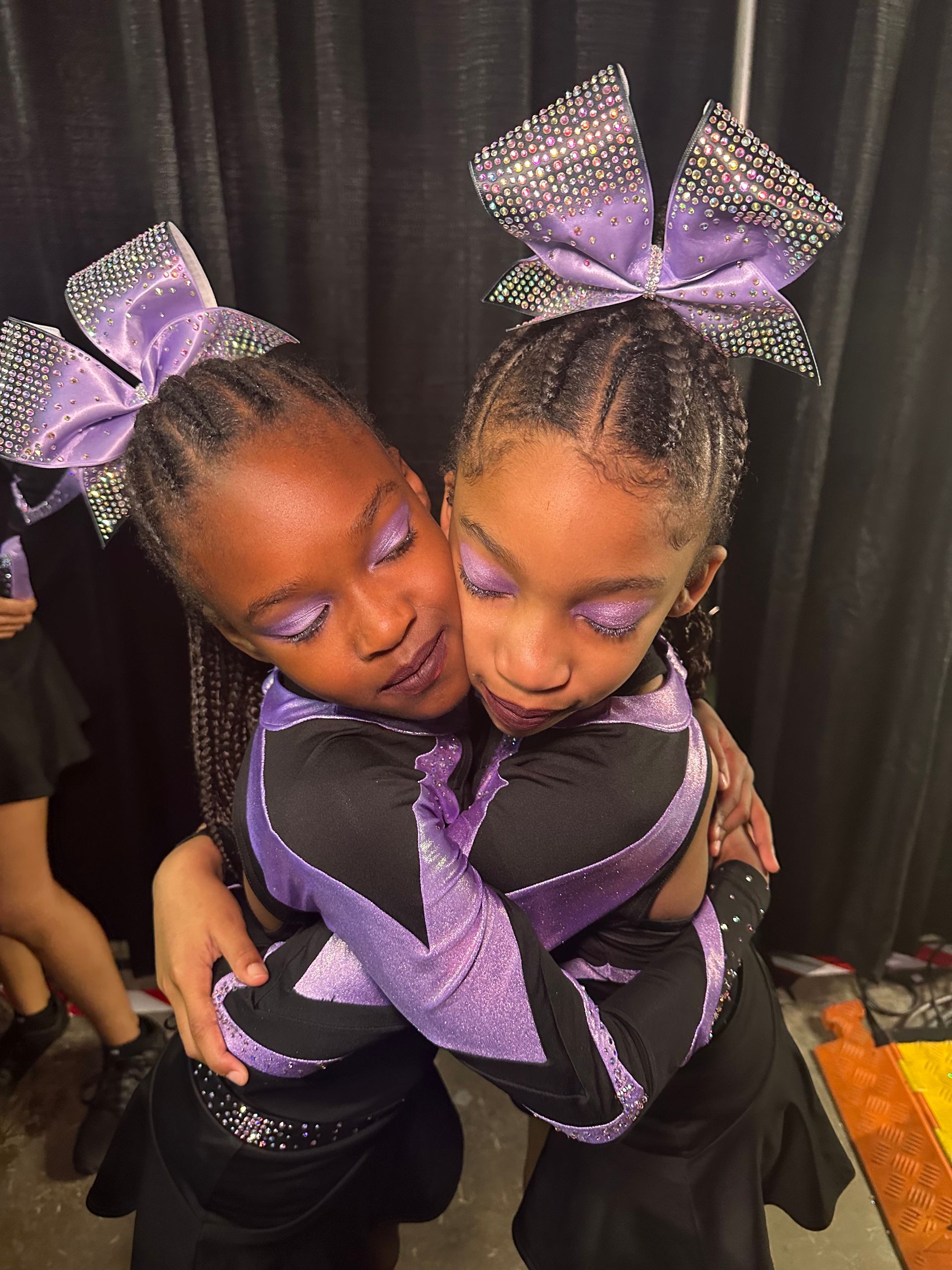 Two cheerleaders are hugging each other in front of a black curtain.