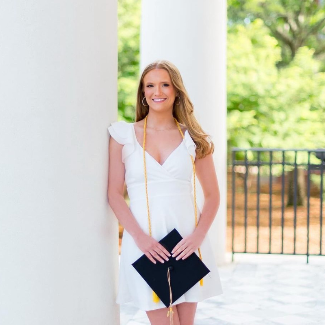 A woman in a white dress is holding a graduation cap and smiling.