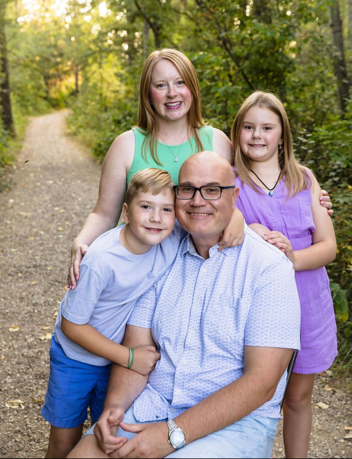 Family of four smiling on a wooded path. Father seated with two children, older daughter behind.