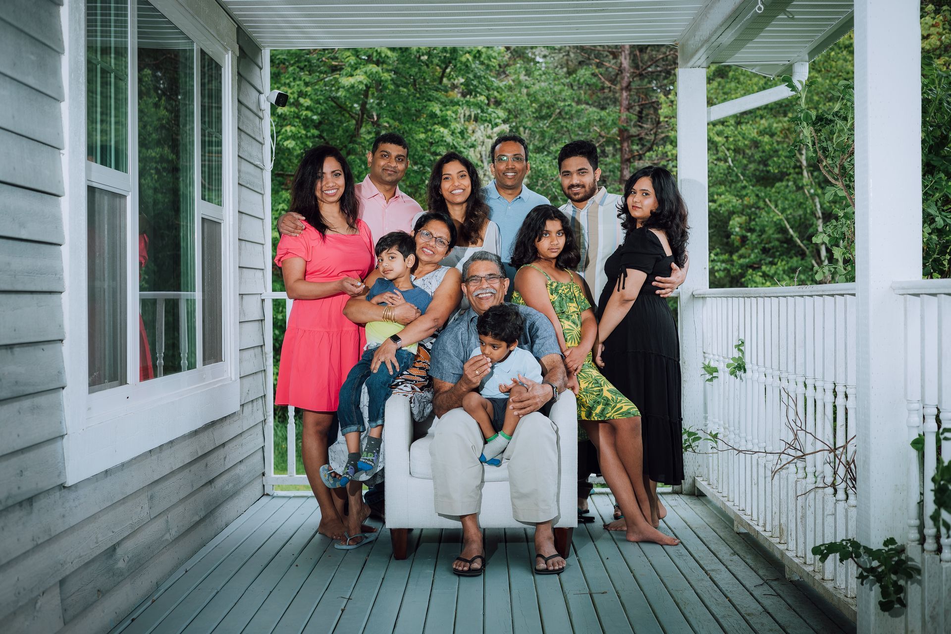 Family group portrait on a porch with green foliage in background. People smiling, seated and standing.