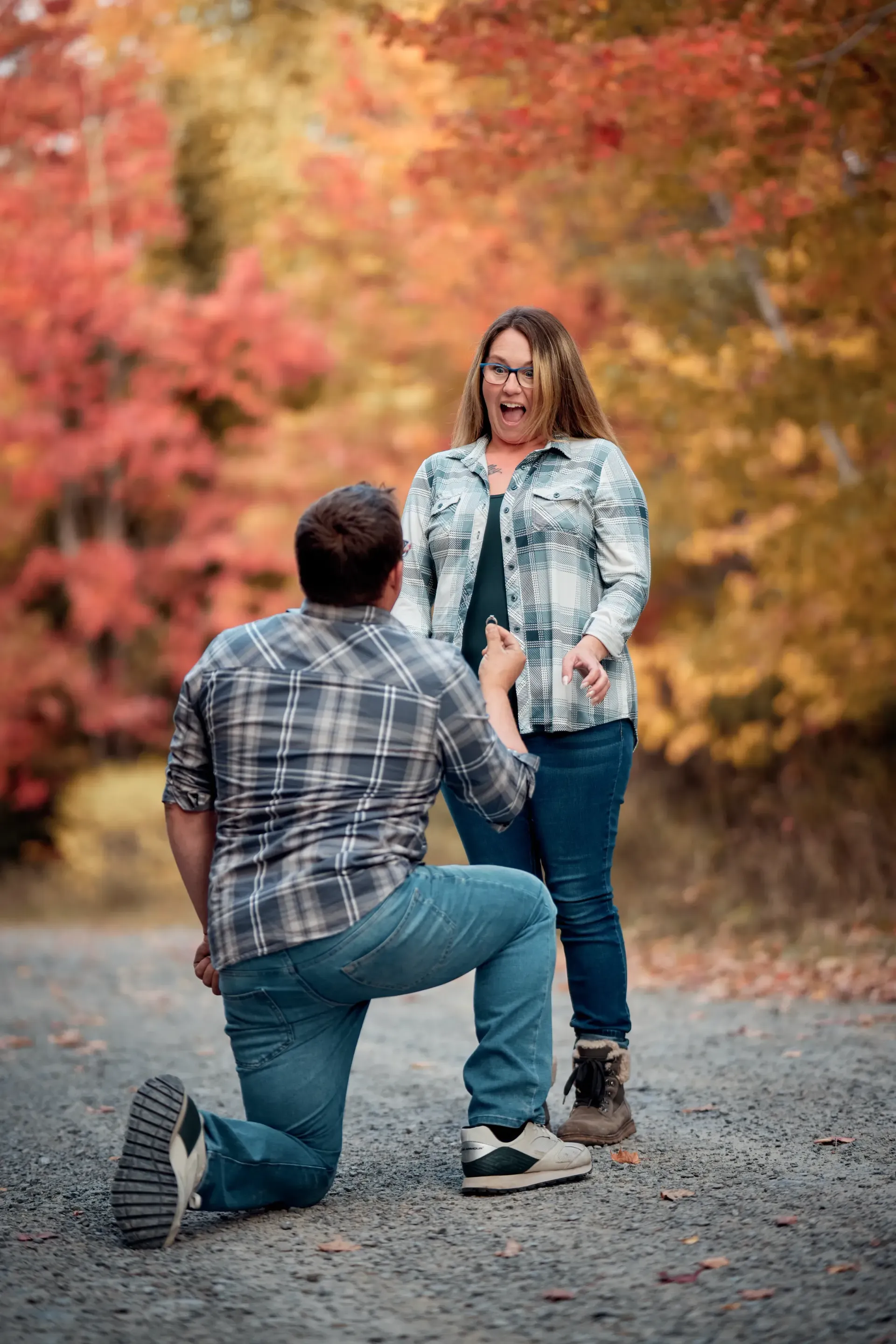 Man kneeling proposing to woman on a path with fall foliage. Woman is surprised, hands up.