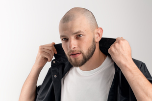 Man with shaved head adjusting black jacket collar over a white shirt.