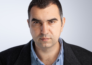 Man with dark hair and a serious expression, wearing a blue plaid shirt and a dark blazer.