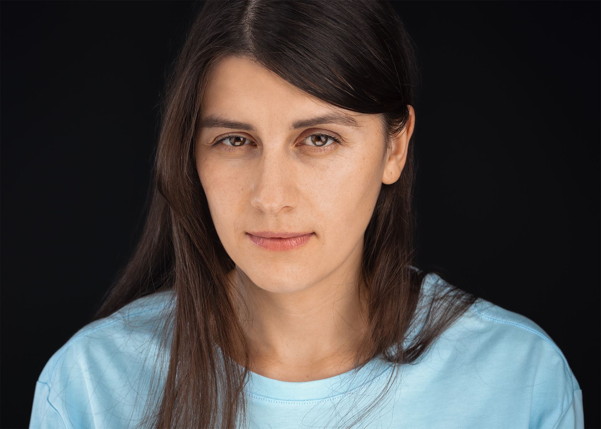 Woman with dark hair in a light blue shirt; neutral expression, black background.