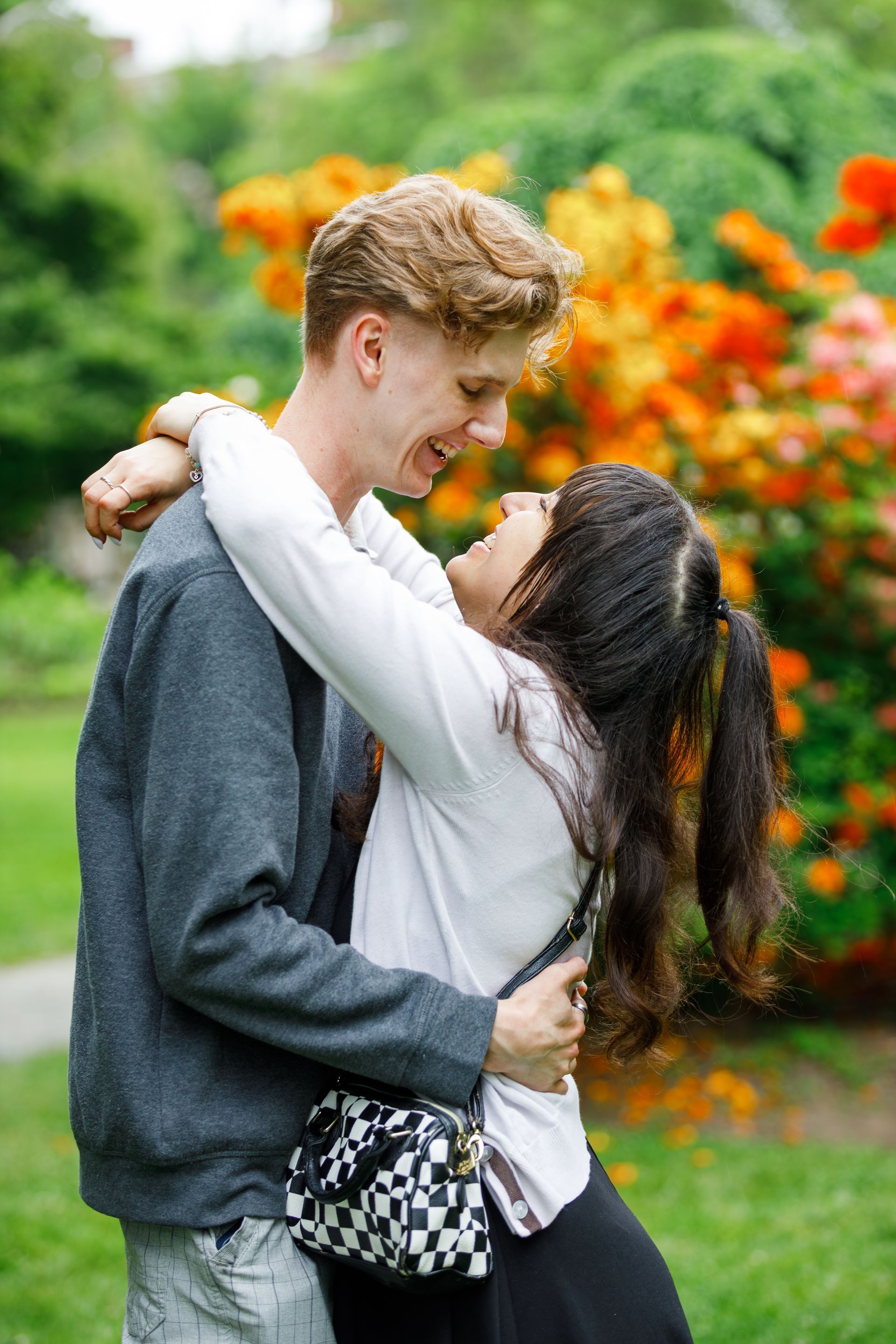 Couple embracing, laughing, outdoors near orange flowers. Man in gray sweater, woman in white shirt.