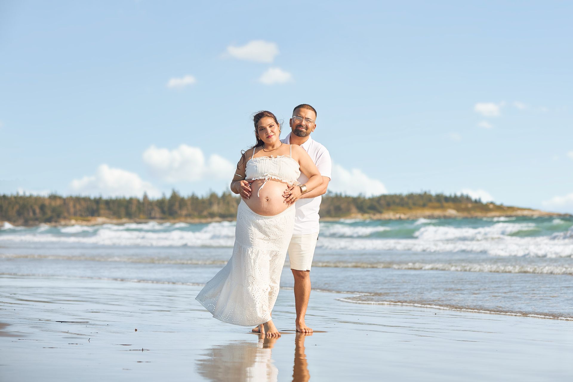 Couple embracing on a beach, woman in white gown cradling her pregnant belly, man with arms around her.