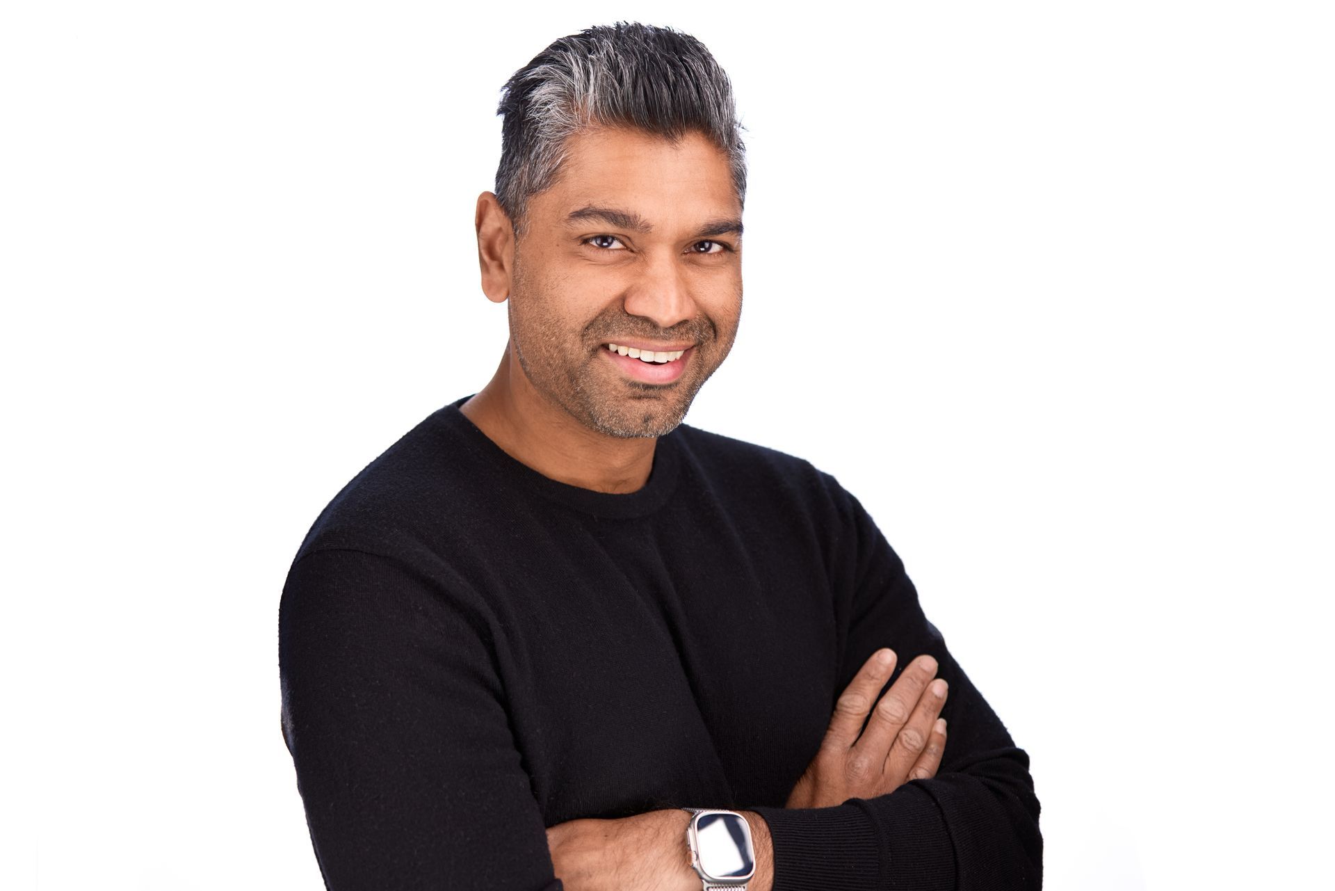 A smiling person with short, grey-streaked dark hair, wearing a black long-sleeved shirt with arms crossed.