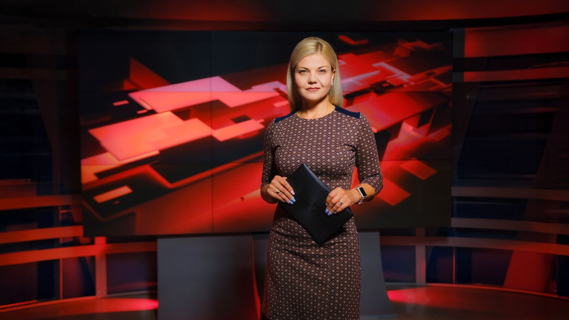 Woman in a dress standing in a news studio holding a tablet, with a red screen background.
