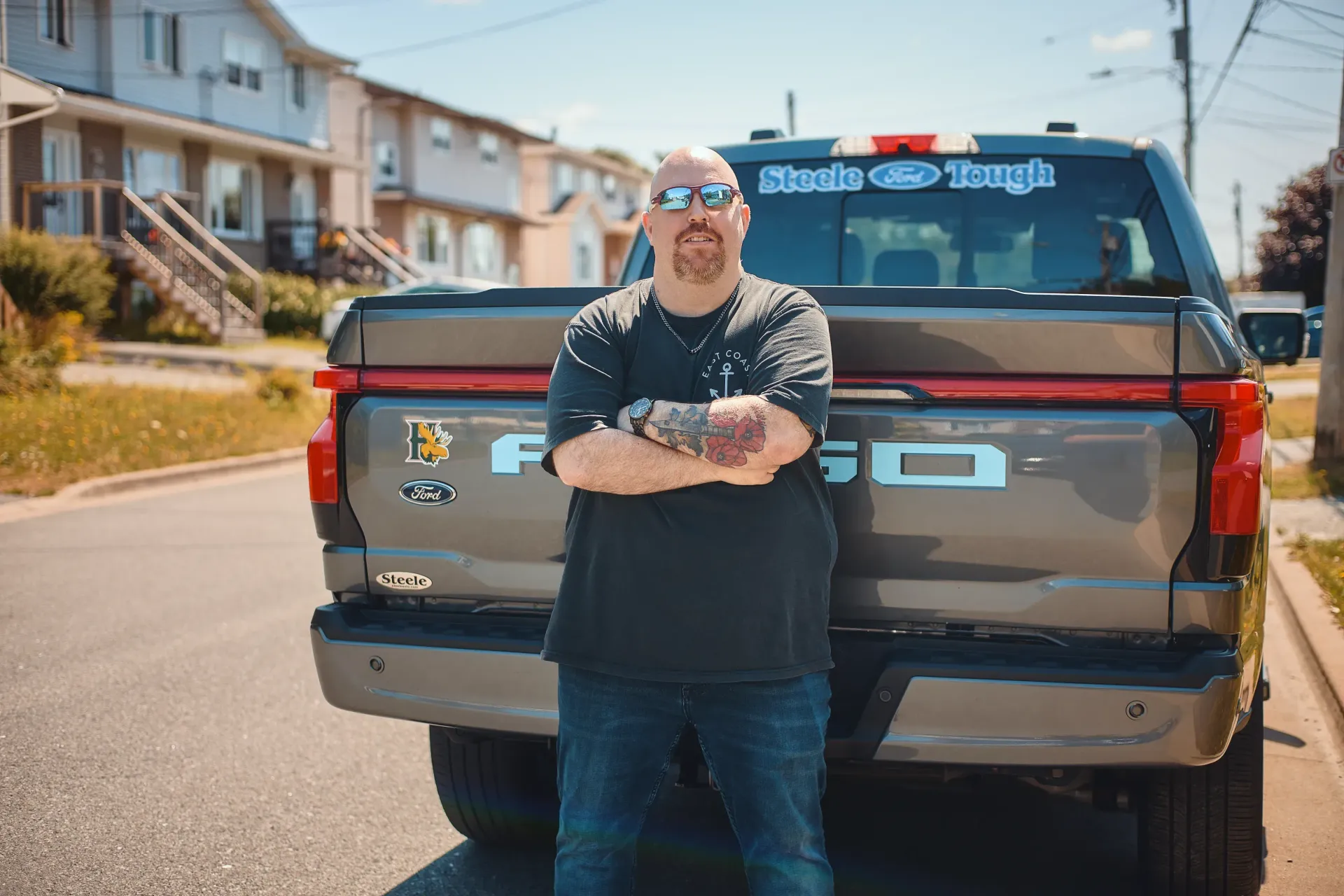 Man with tattoos and sunglasses stands in front of a gray Ford F-150 truck on a sunny residential street.