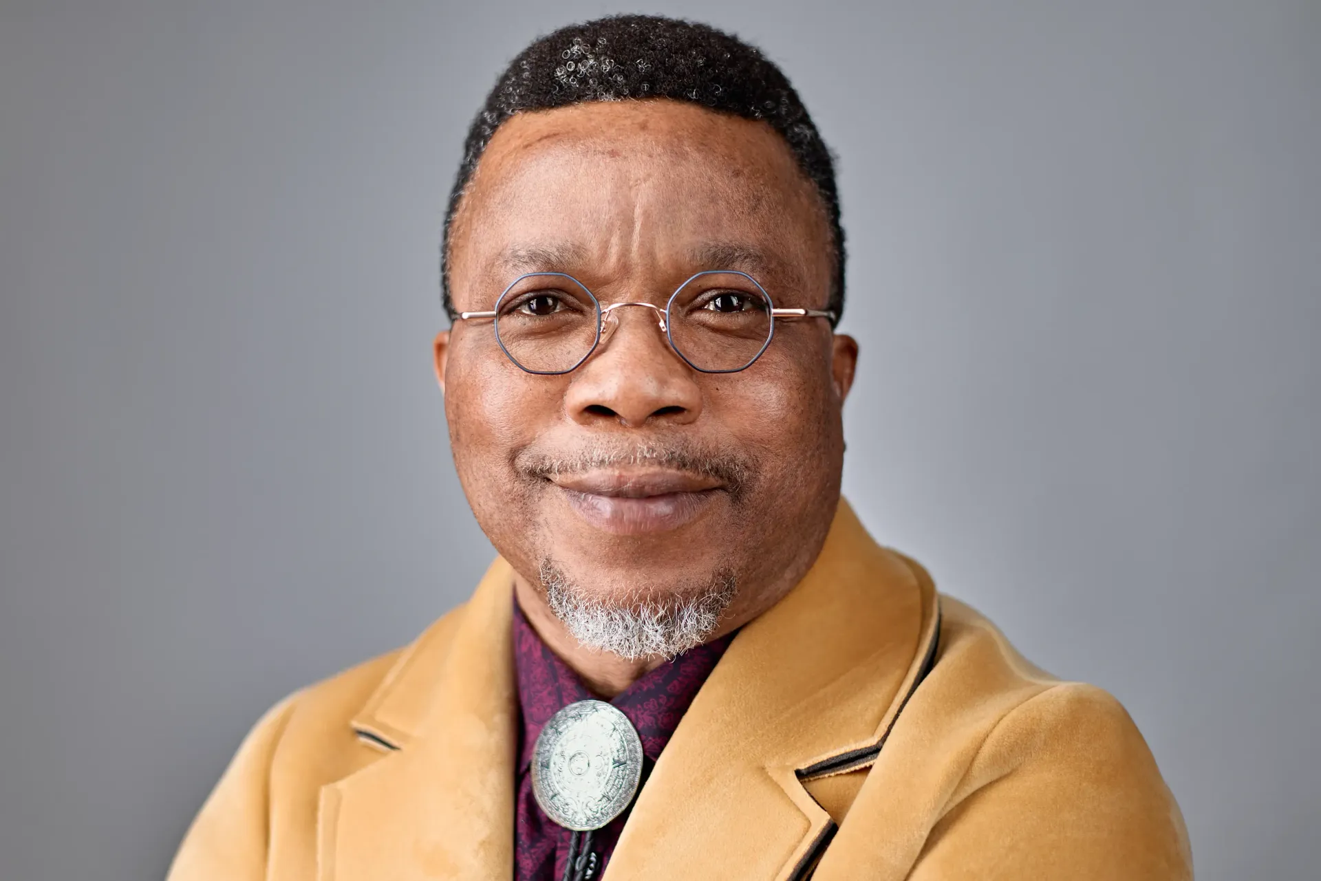 A portrait of a person with short hair, wearing glasses, a dark shirt, a bolo tie, and a tan blazer against a gray backdrop.
