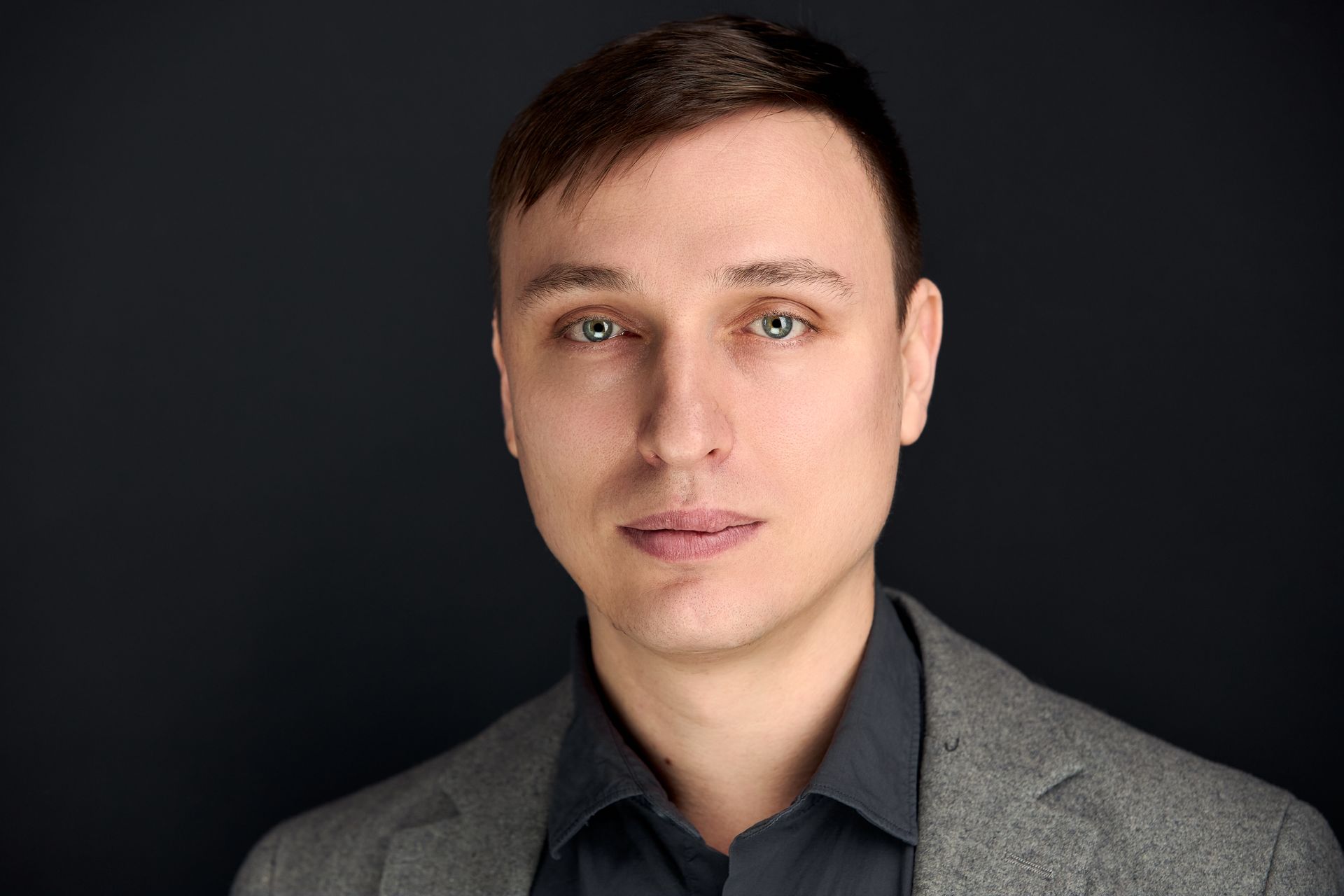 Man with light skin wearing a grey blazer and dark shirt, looking at the viewer.