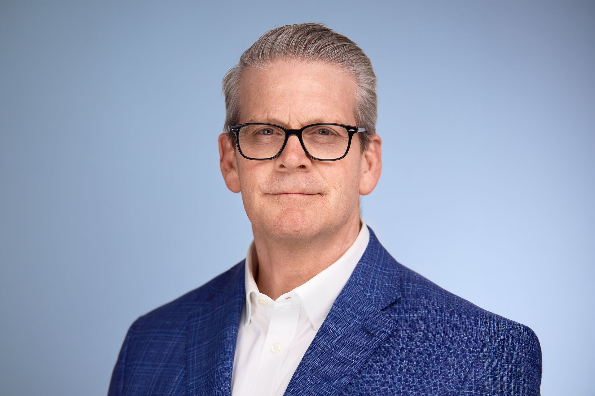 A person wearing glasses and a blue textured blazer over a white collared shirt against a light blue background.