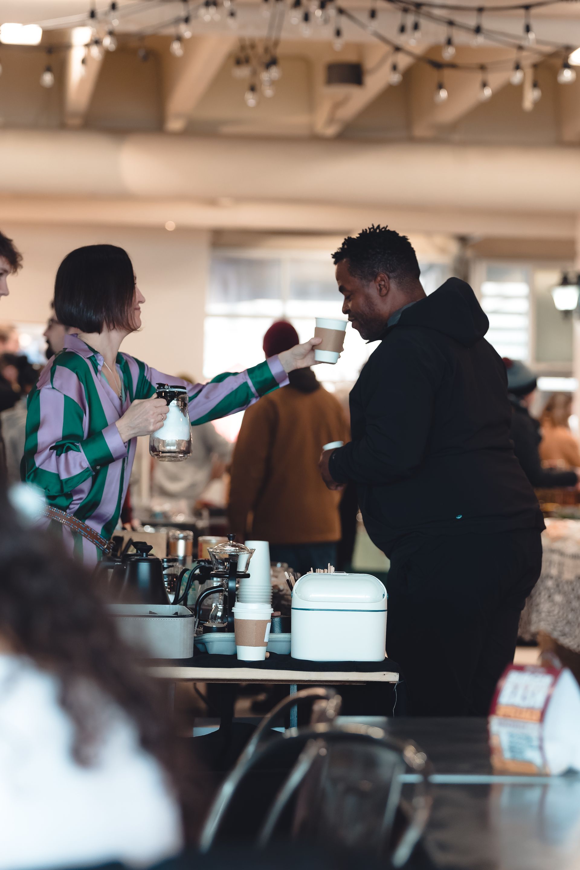 Woman in striped shirt pours coffee for a man in a black hoodie at a cafe counter.