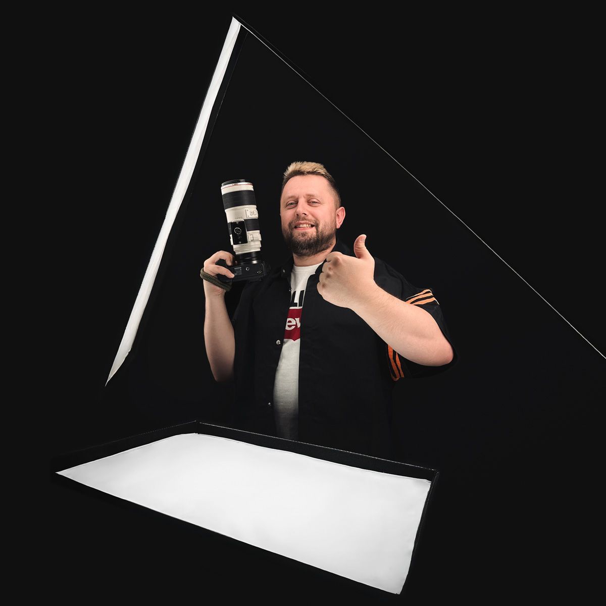 Man holding camera gives thumbs up, lit by a triangle of light in front of a black background.