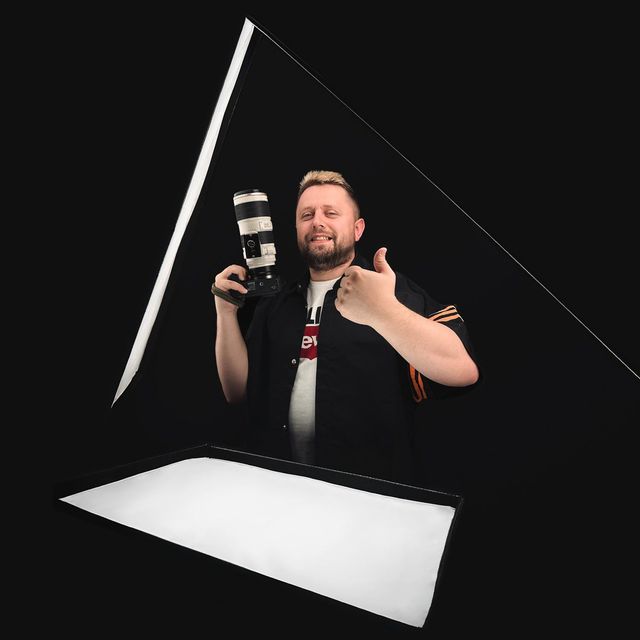 Man holding camera gives thumbs up, lit by a triangle of light in front of a black background.