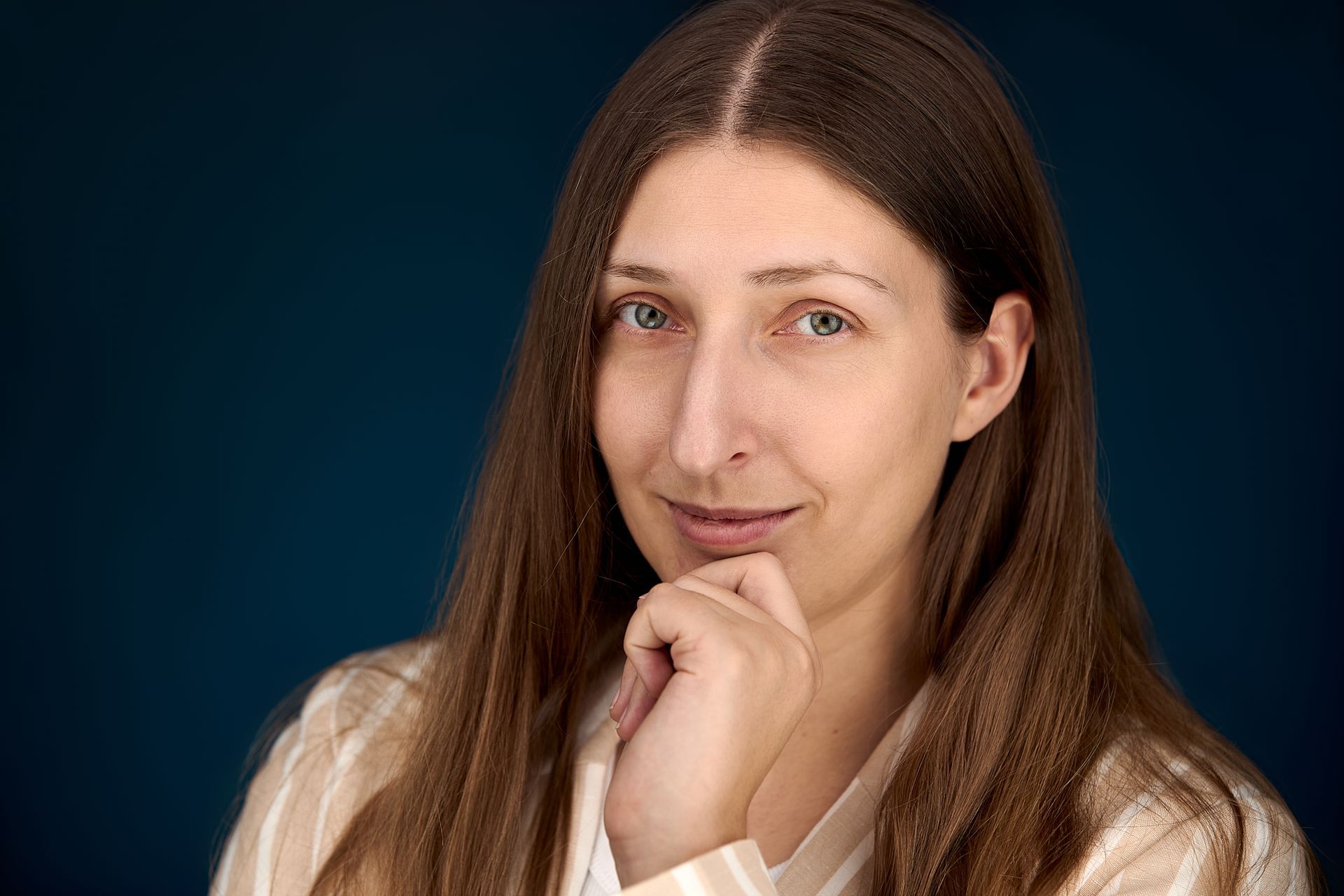 Woman with long brown hair, resting chin on hand, smiling, against a dark blue background.