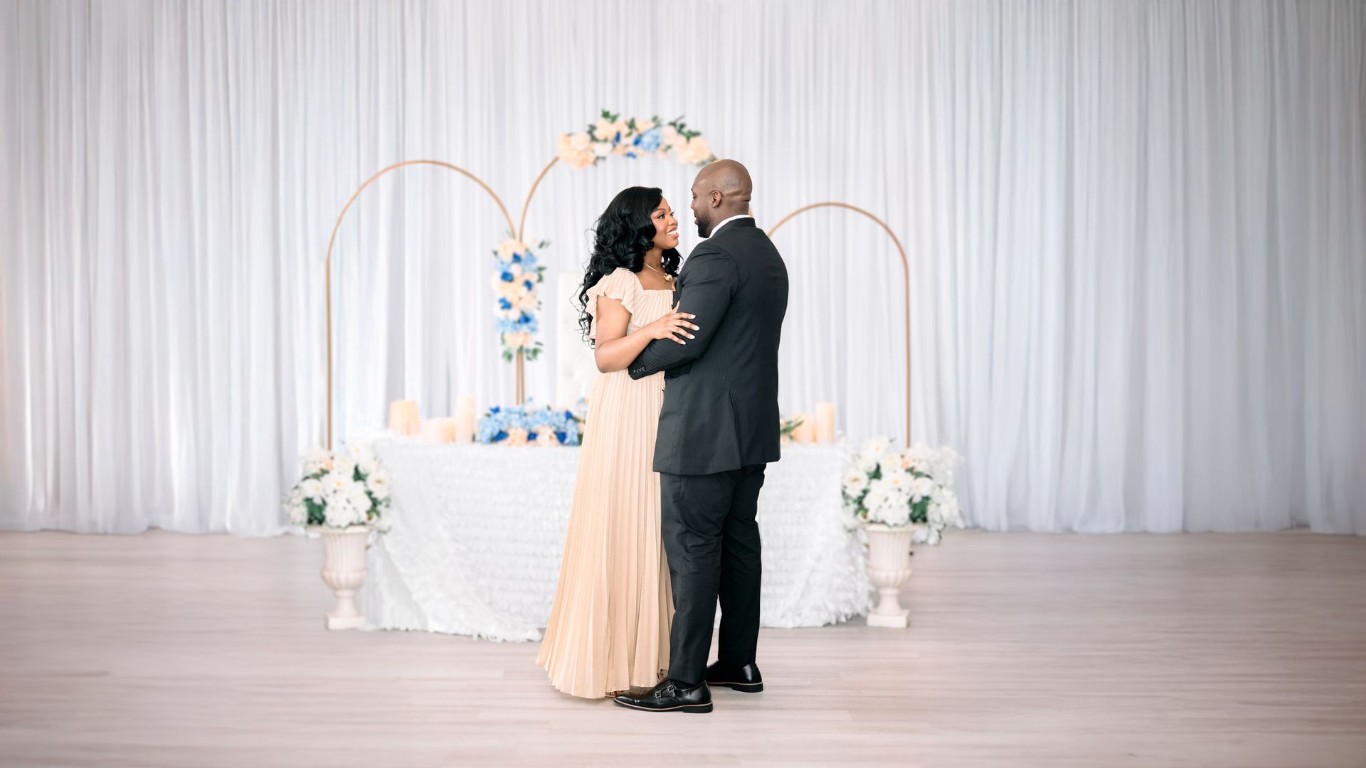 Couple embraces in front of floral-decorated arch and table with draped white backdrop.