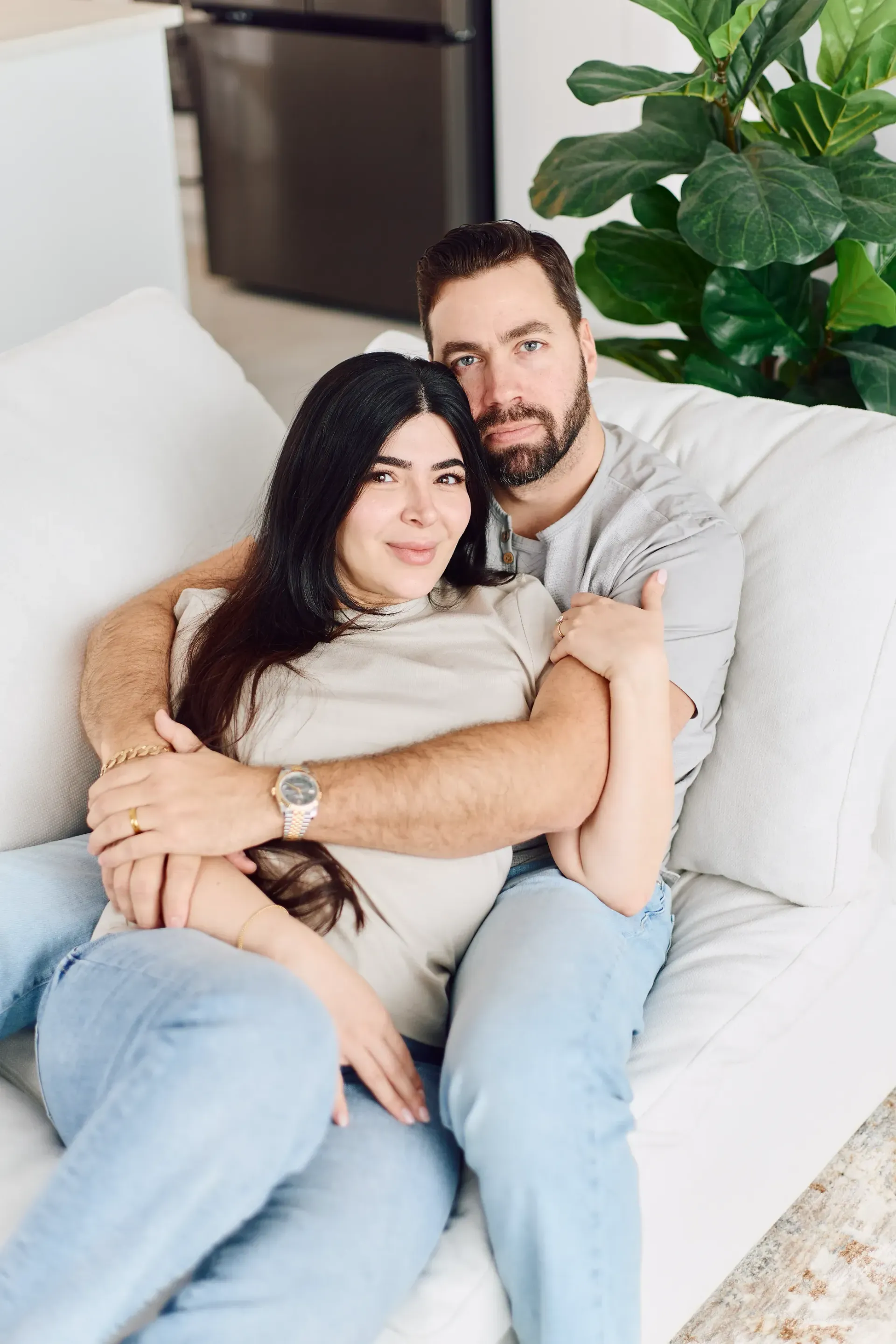 Couple cuddling on a white couch. Man with beard wraps arms around woman. Brightly lit living room.