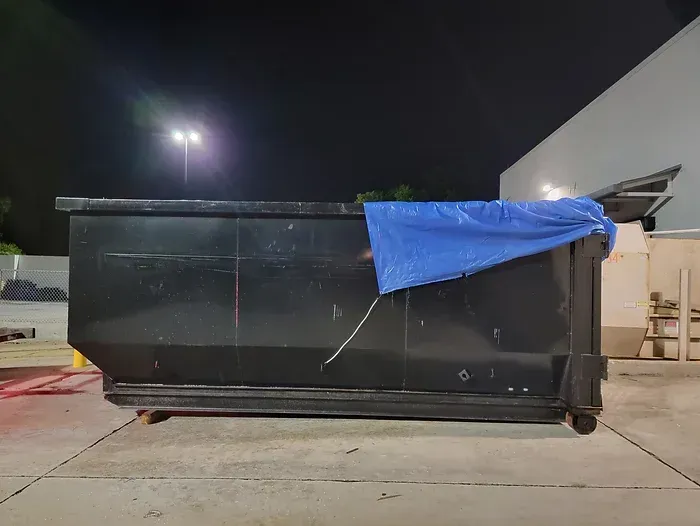 A dumpster with a blue tarp on top of it is parked in a parking lot at night.