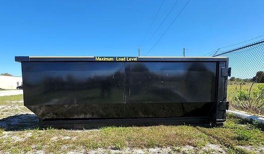 A large black dumpster is sitting in a grassy field next to a chain link fence.