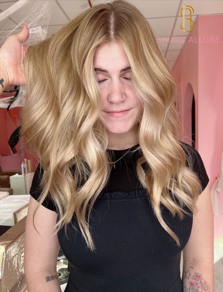 A person with blonde wavy hair posing against a solid pink background, holding their hands up near their cheeks.
