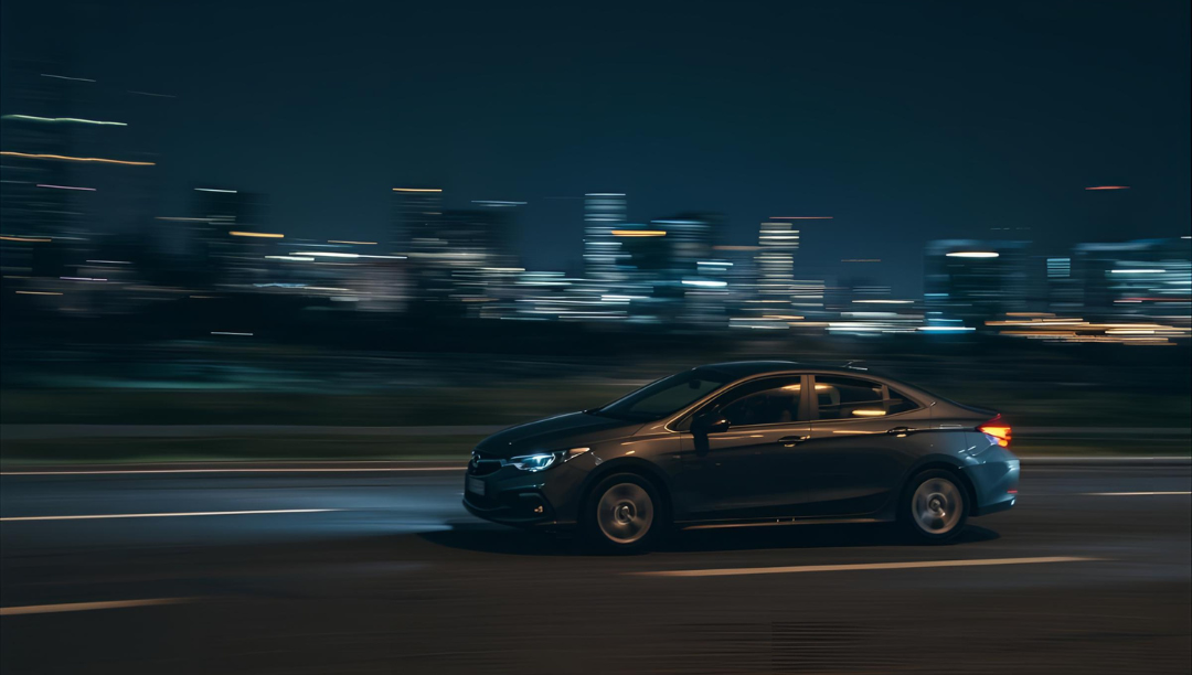Dark car driving fast on a road at night, city lights blurred in the background.