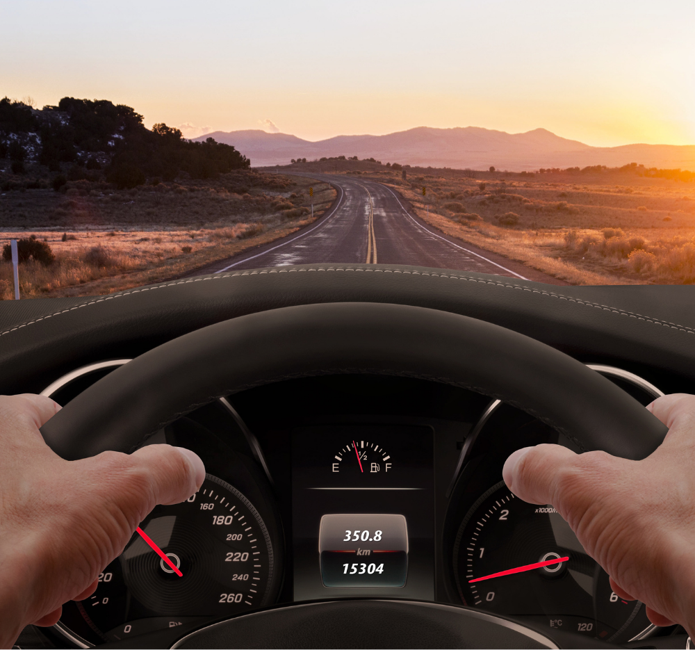 Hands gripping a steering wheel, driving on a road towards a sunset. Dashboard visible, with speed and mileage displayed.