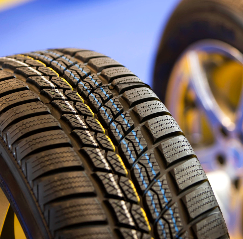 Close-up of a winter tire with deep tread, partially obscuring a shiny silver wheel; blue and yellow background.