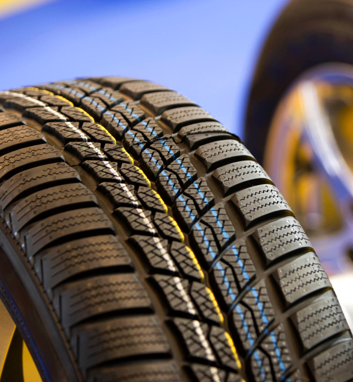 Close-up of a winter tire with deep tread, partially obscuring a shiny silver wheel; blue and yellow background.