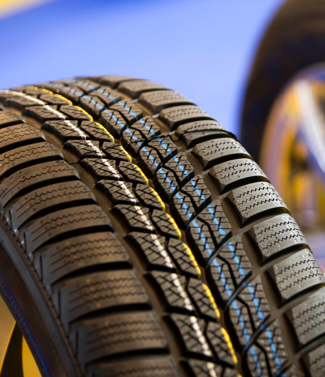 Close-up of a winter tire with deep tread, partially obscuring a shiny silver wheel; blue and yellow background.