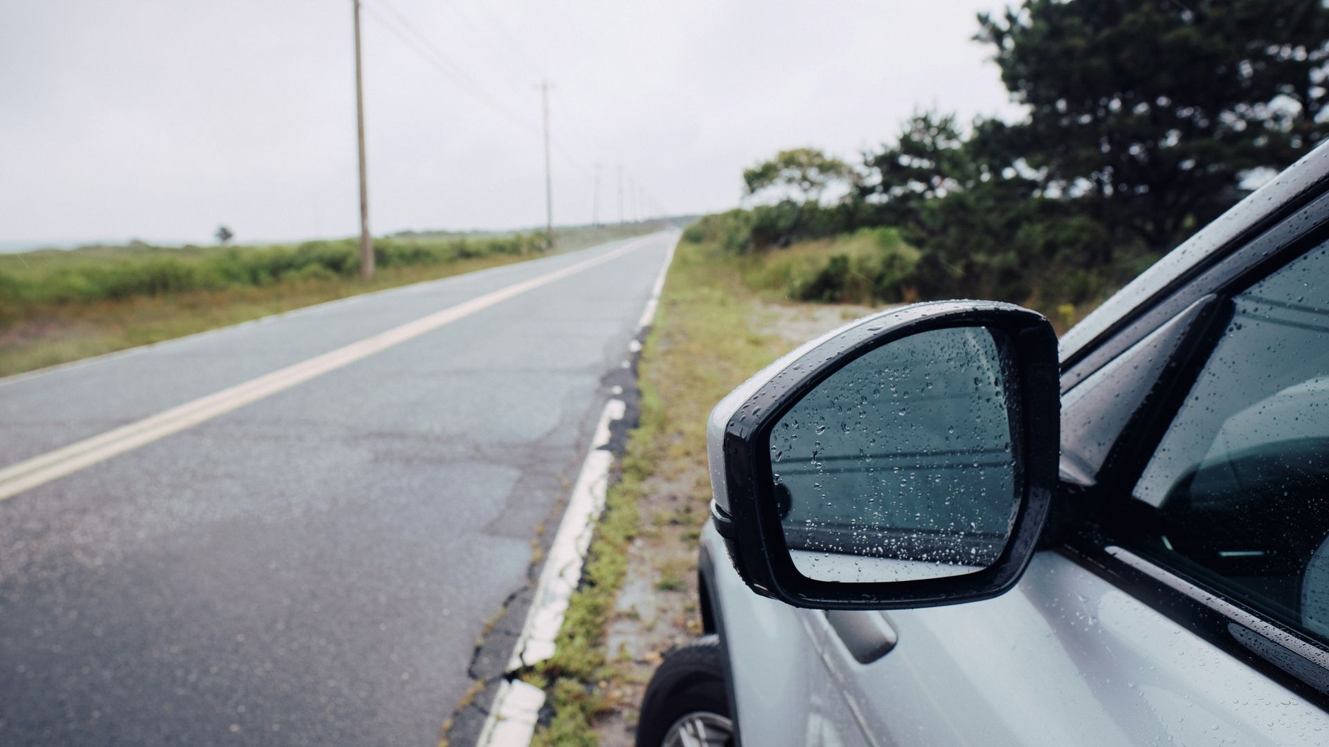 Rainy roadside beside a silver car on an empty wet road with cloudy sky
