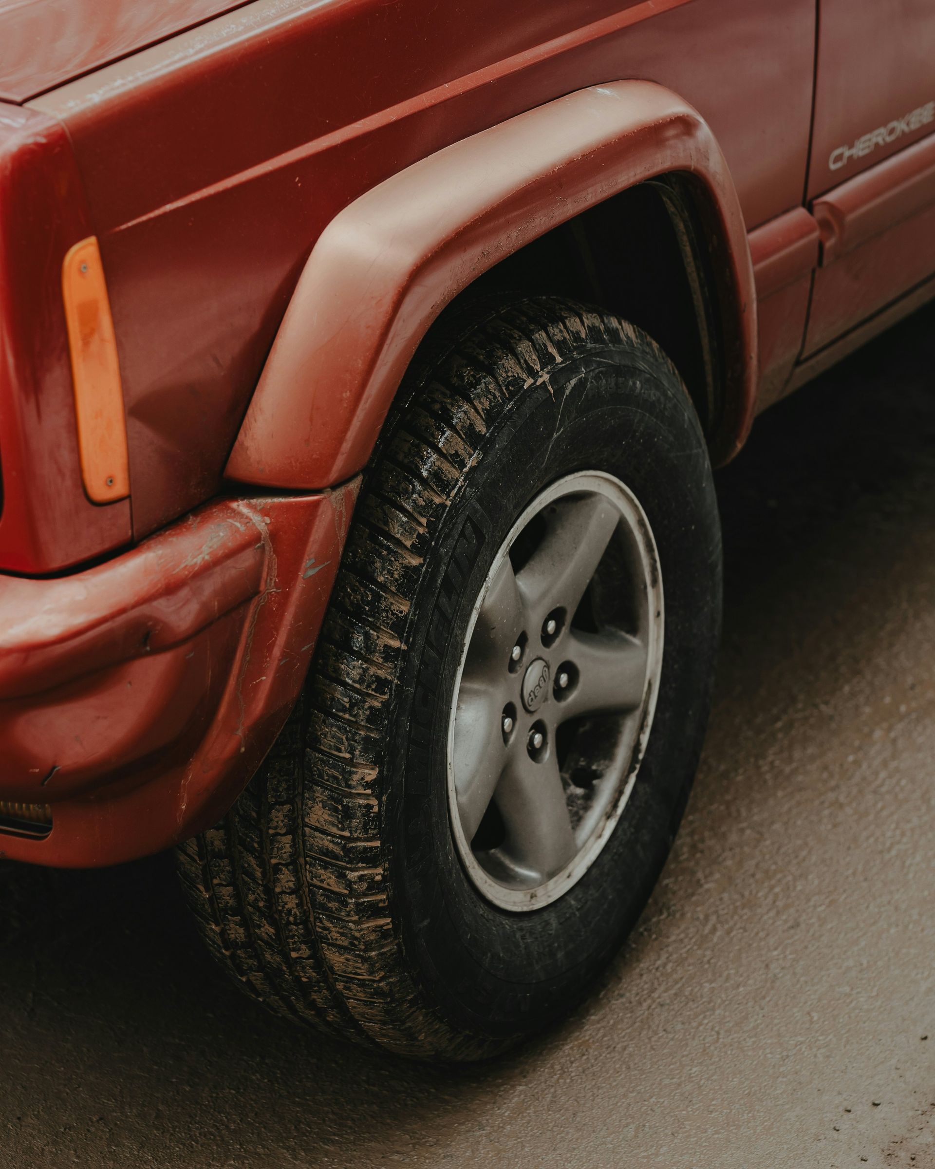Close-up of the front driver-side wheel and fender of a red Jeep Cherokee parked on a gray, paved surface.