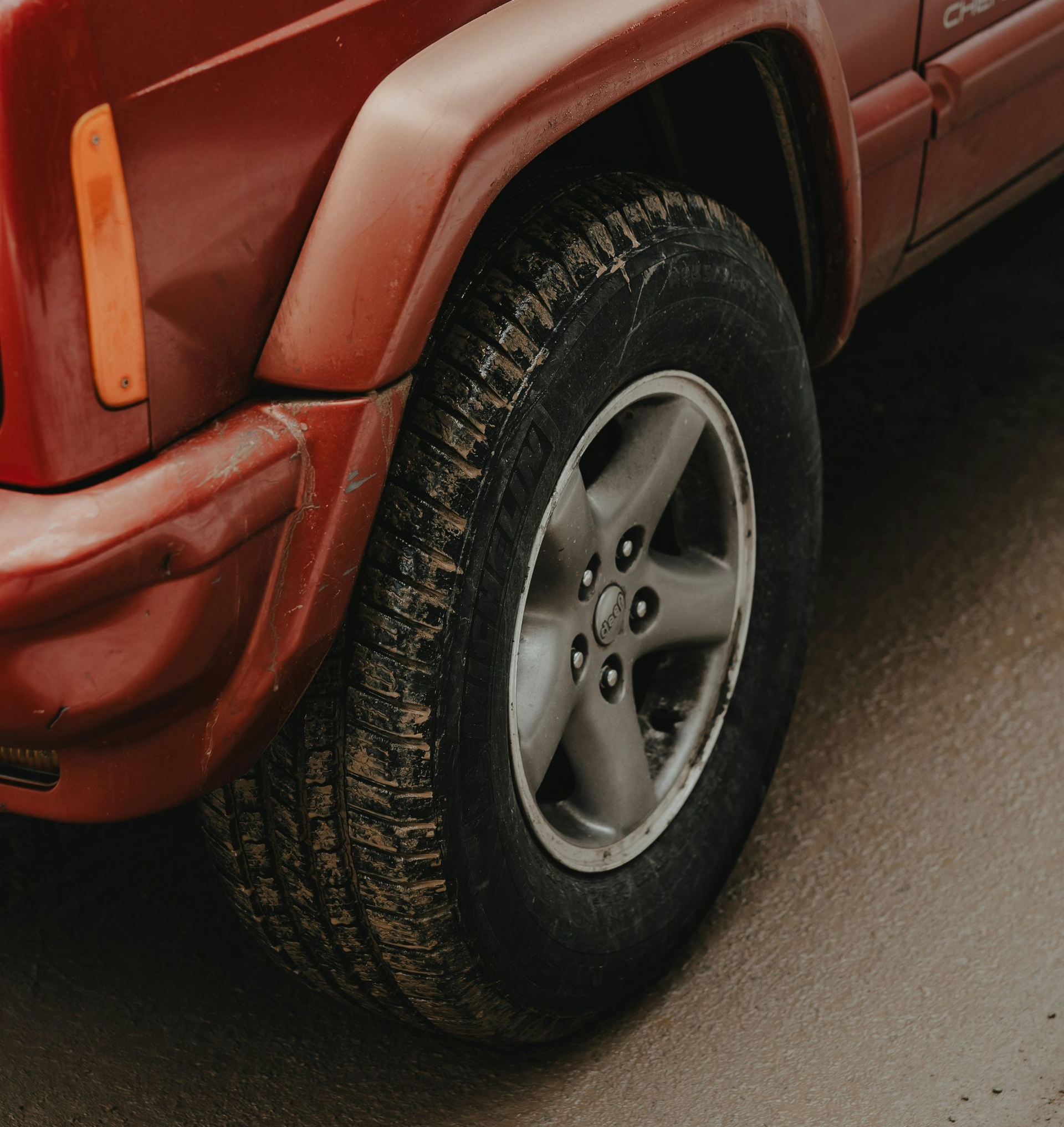 Close-up of the front driver-side wheel and fender of a red Jeep Cherokee parked on a gray, paved surface.
