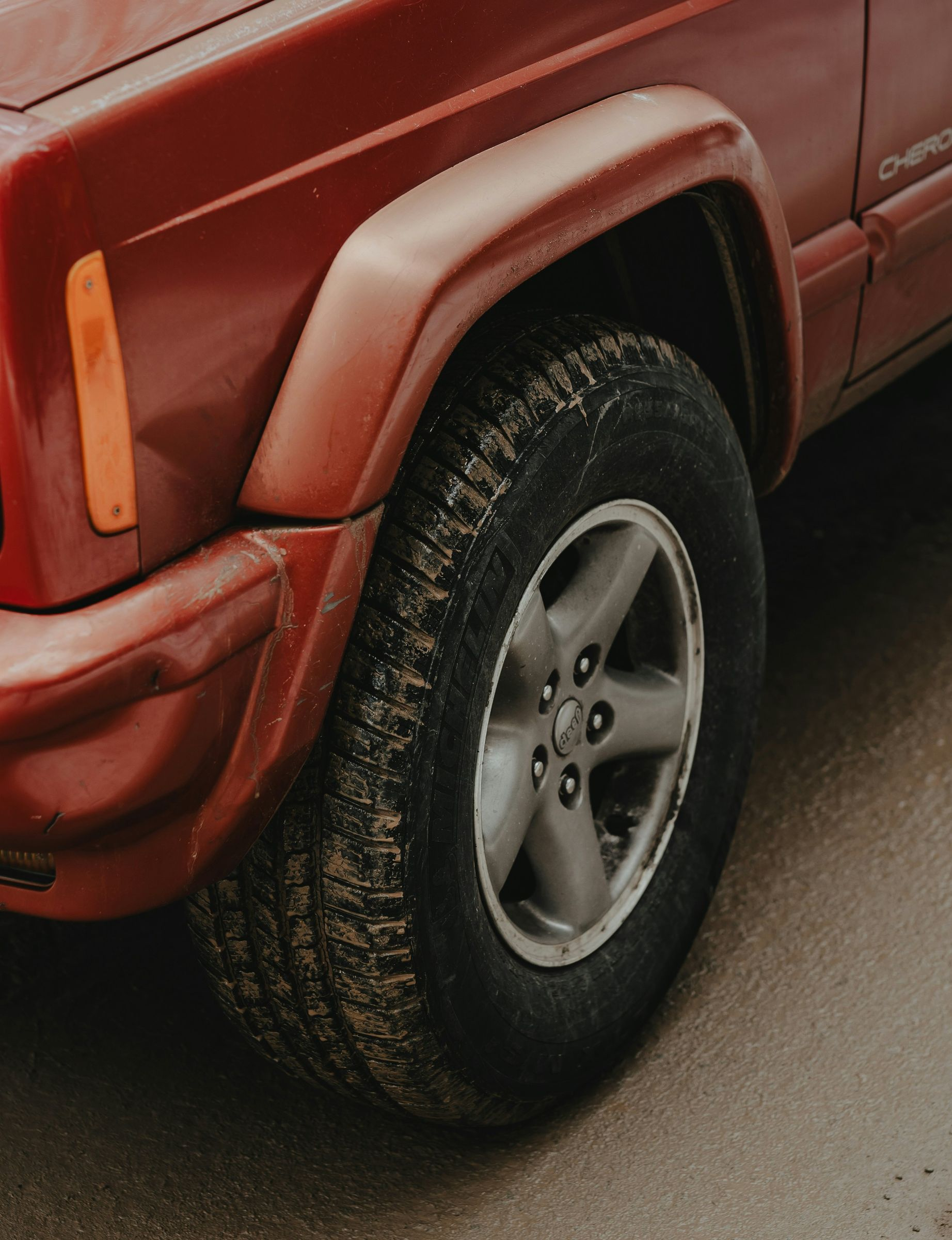 Close-up of the front driver-side wheel and fender of a red Jeep Cherokee parked on a gray, paved surface.