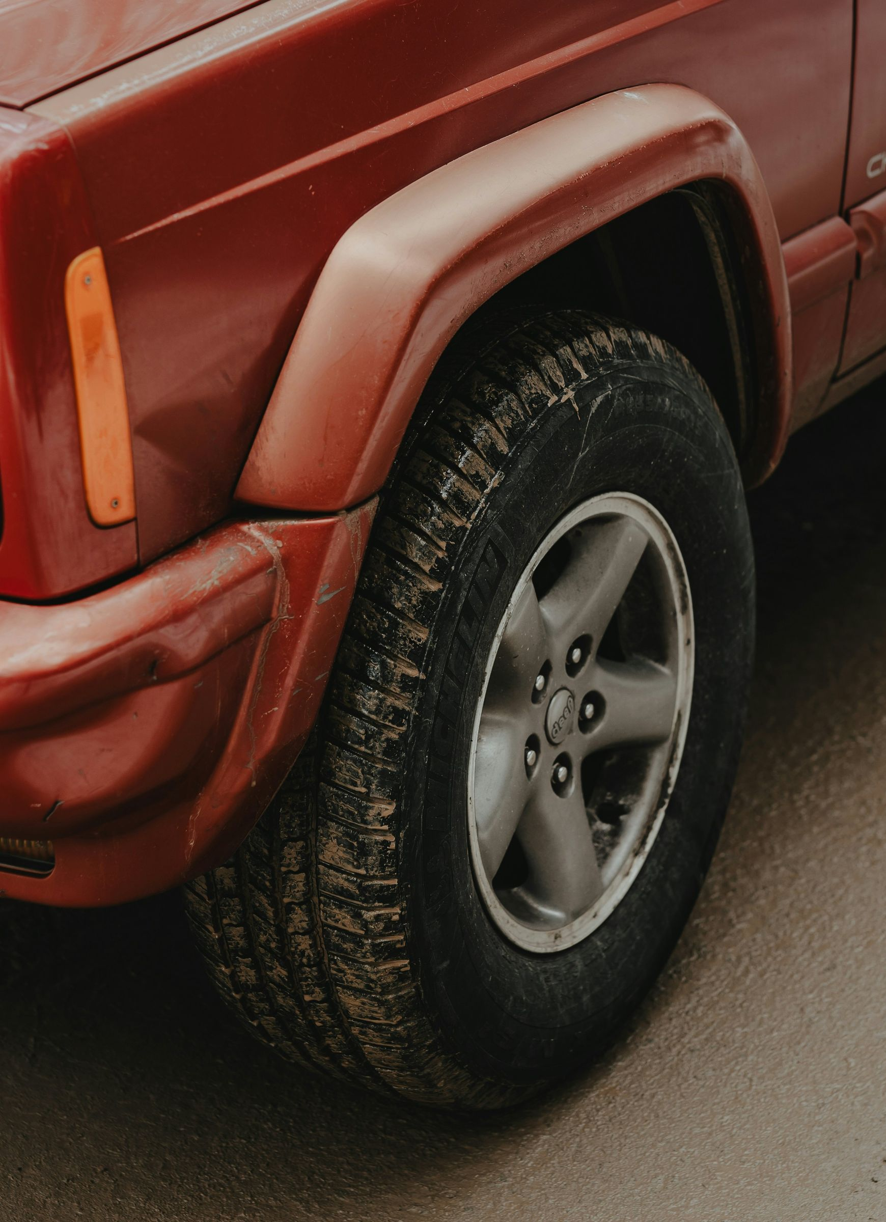 Close-up of the front driver-side wheel and fender of a red Jeep Cherokee parked on a gray, paved surface.