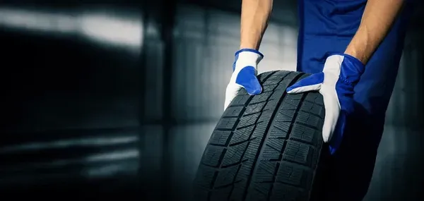 Mechanic in blue uniform and gloves holding a tire in a dark garage.