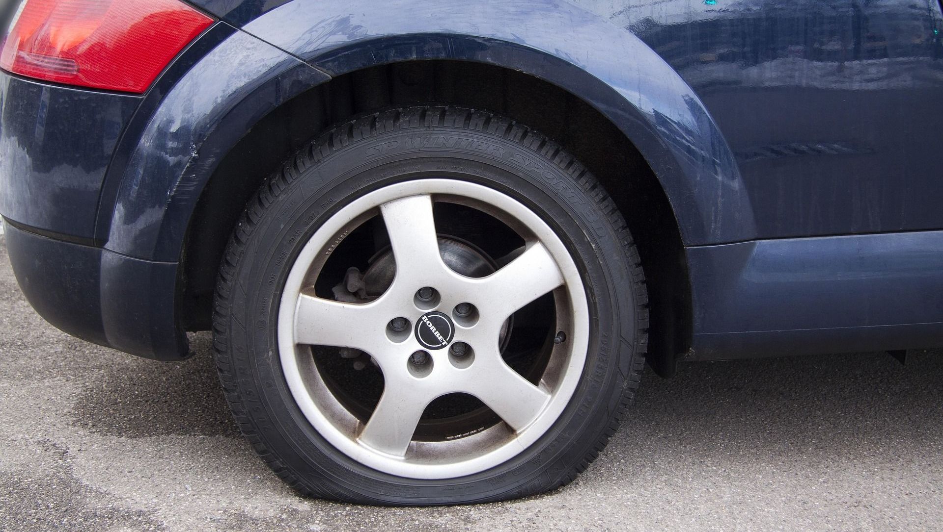 A flat tire on a blue car. The tire is deflated, and the car is parked on a gray surface.