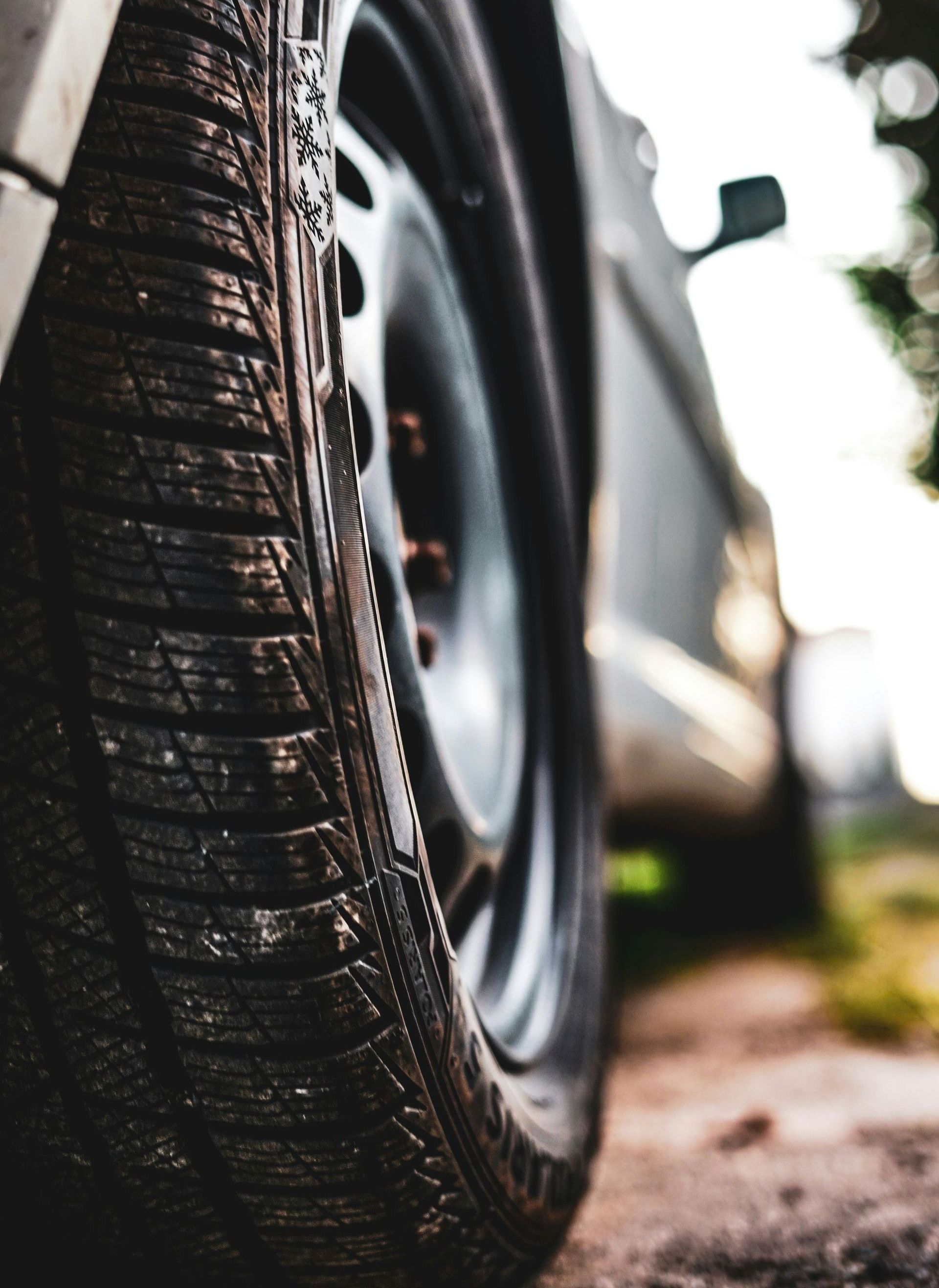 Close-up of a car tire on a dusty road, showing tread detail. Gray car body in background.