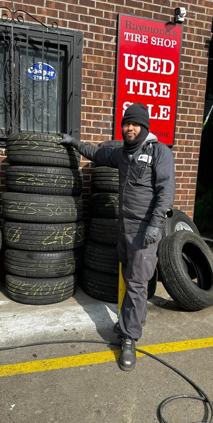 Man stands by tires stacked outside a used tire shop. Shop has a red sign, brick building.