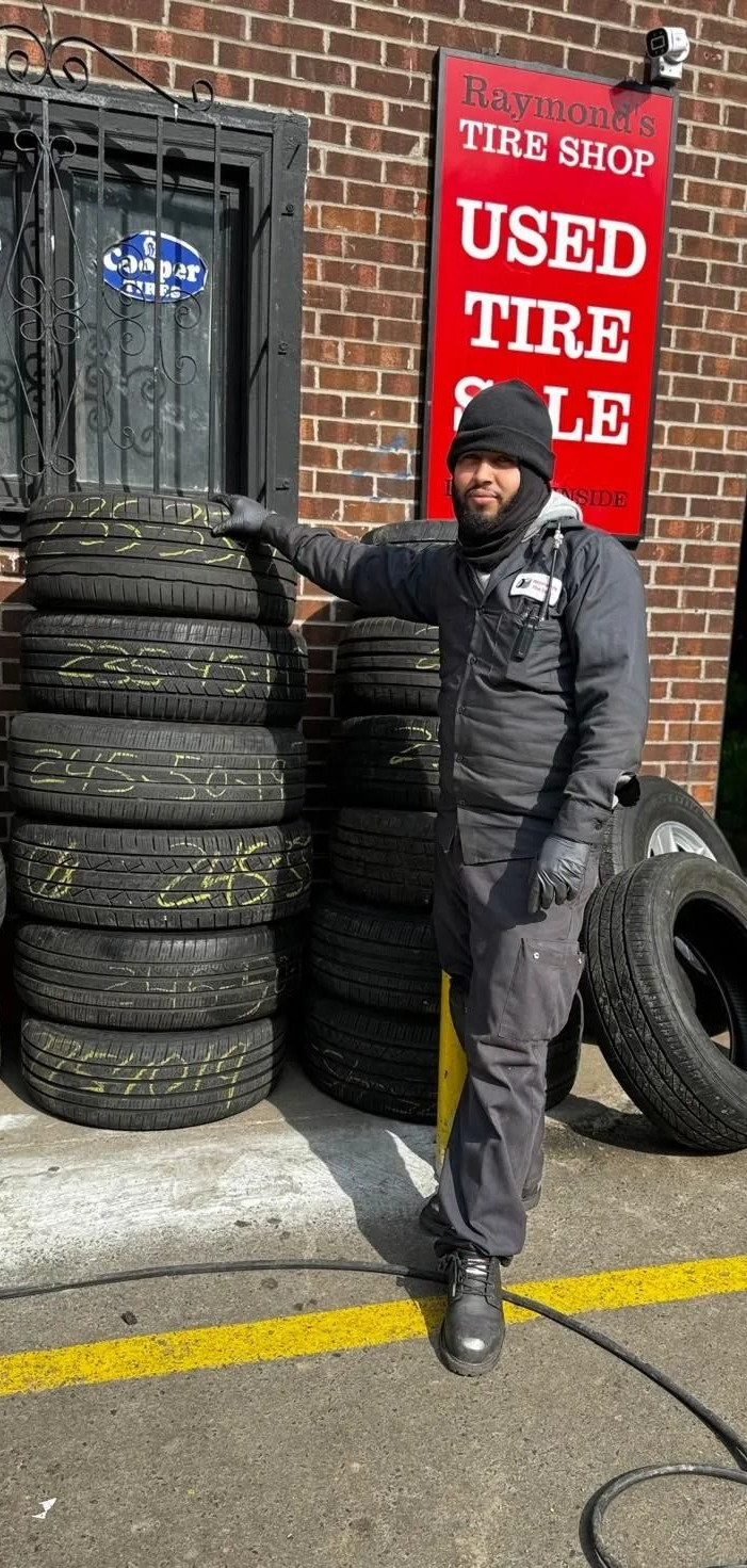 Man stands by tires stacked outside a used tire shop. Shop has a red sign, brick building.