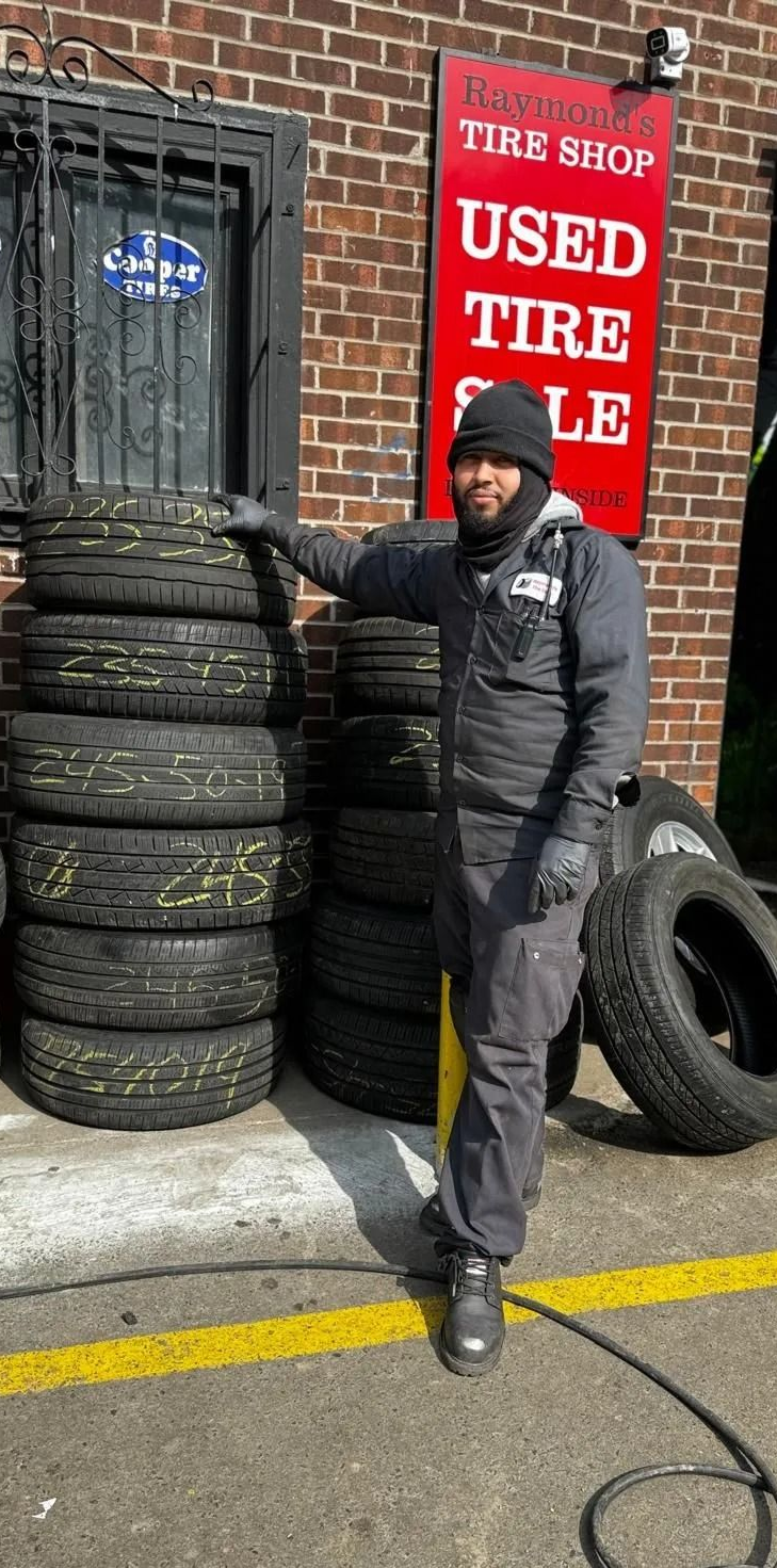 Man stands by tires stacked outside a used tire shop. Shop has a red sign, brick building.