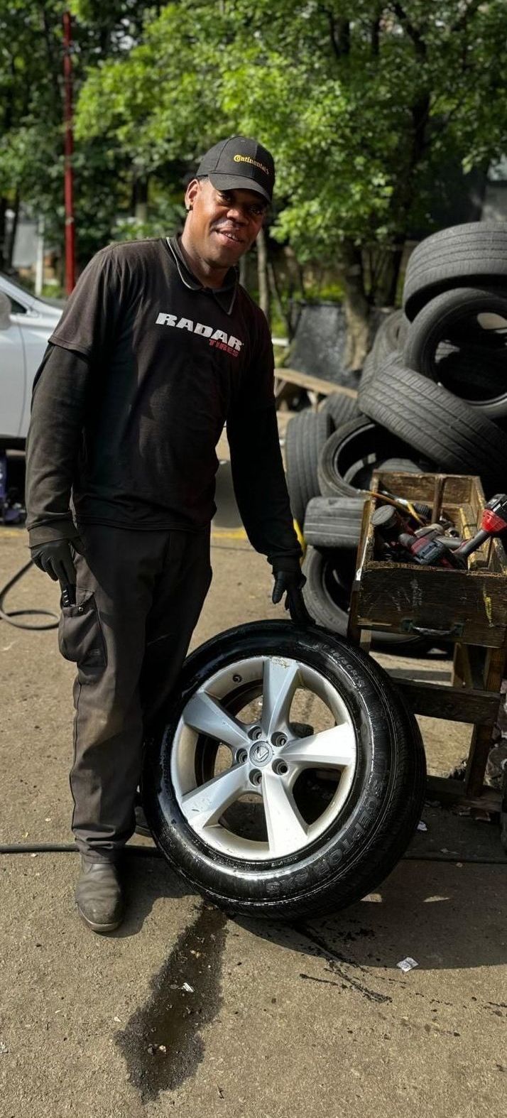 A mechanic, wearing a black cap and gloves, smiles and holds a tire with a silver rim outside.