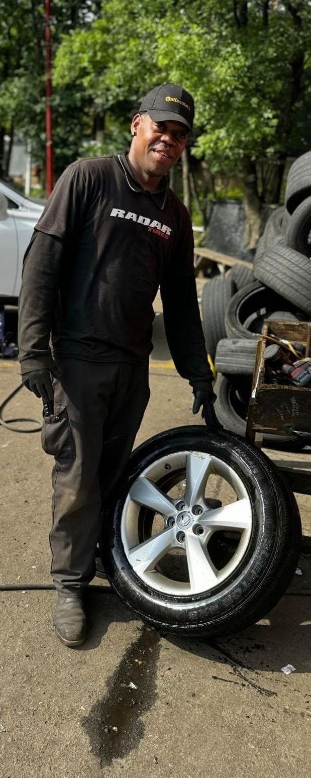 Mechanic holding a tire and smiling in a shop. He wears a cap, black shirt, gloves, and stands near a tire pile.