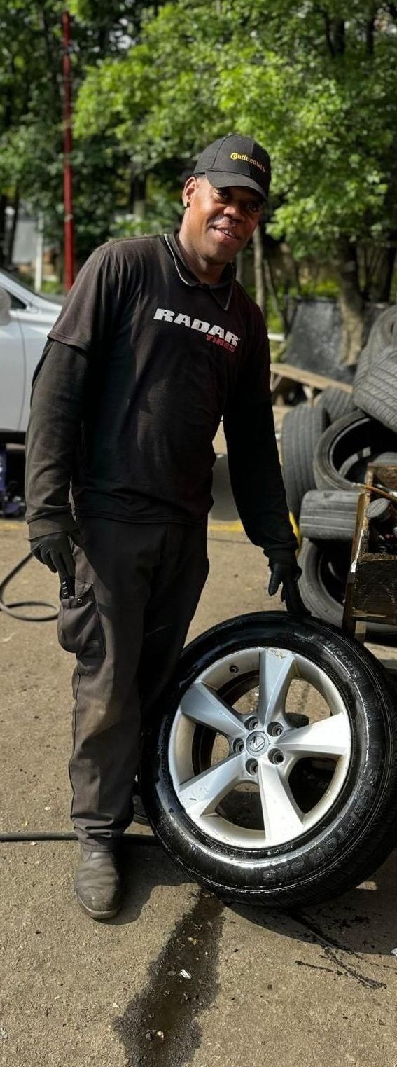 Man in work clothes holds a car tire and rim. He is outside, near a pile of tires.